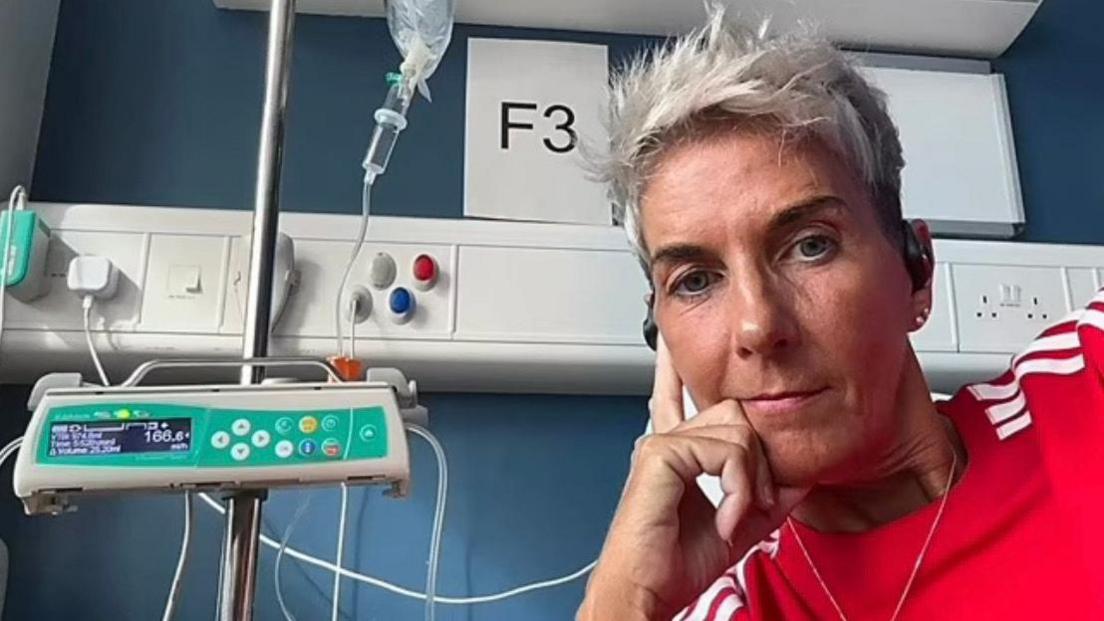 Carmel is looking at the camera and is leaning against her hand. She is in a hospital setting and wires and tubes can be seen beside her. She is wearing a red top with white stripes and has short grey hair. 