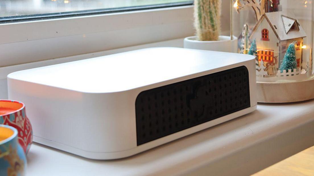 White plastic box (approx the size of a ream of A4 paper) with a black panel on the front sitting on a shelf in a house surrounded by ornaments.