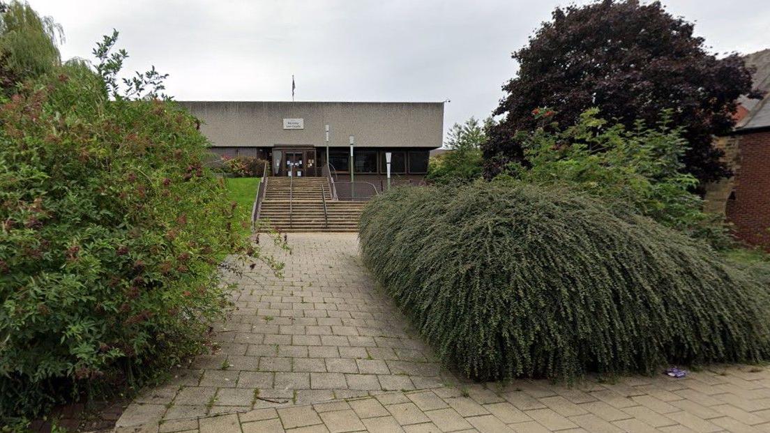 A picture showing Barnsley Magistrates' Court. It is grey coloured building with steps up to the entrance. There are several bushes surrounding the building. 