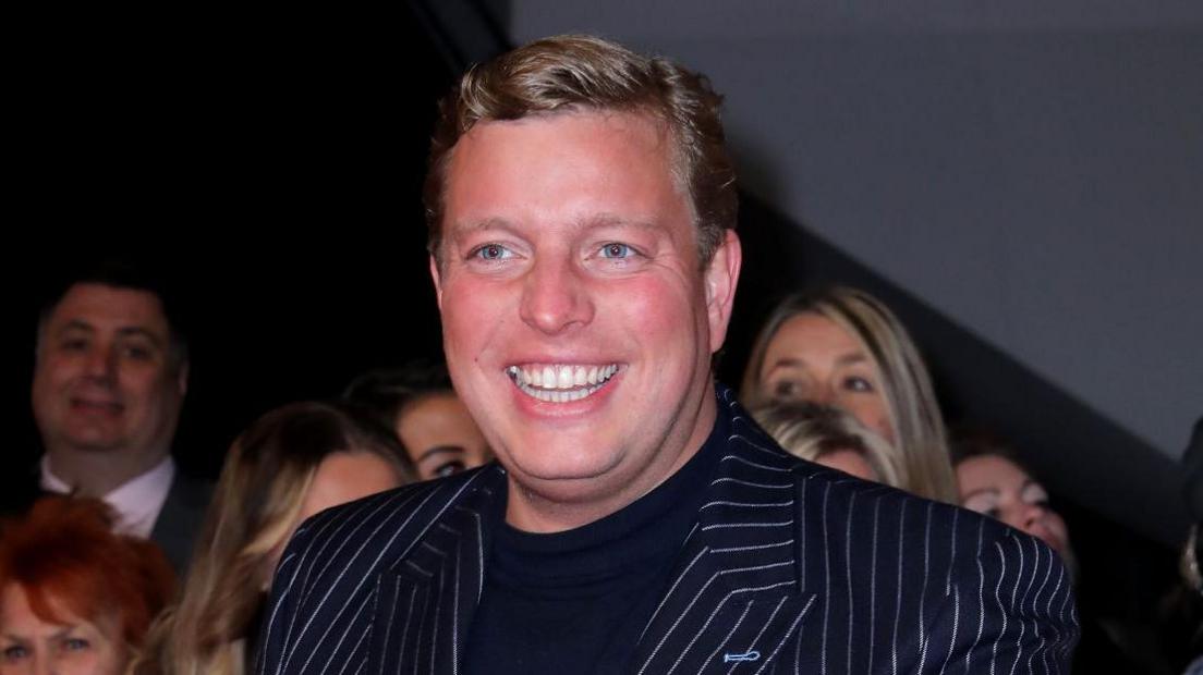 Thomas Skinner smiling in front of a small crowd. He has short blonde hair and is wearing a black T-shirt and black and white striped suit.