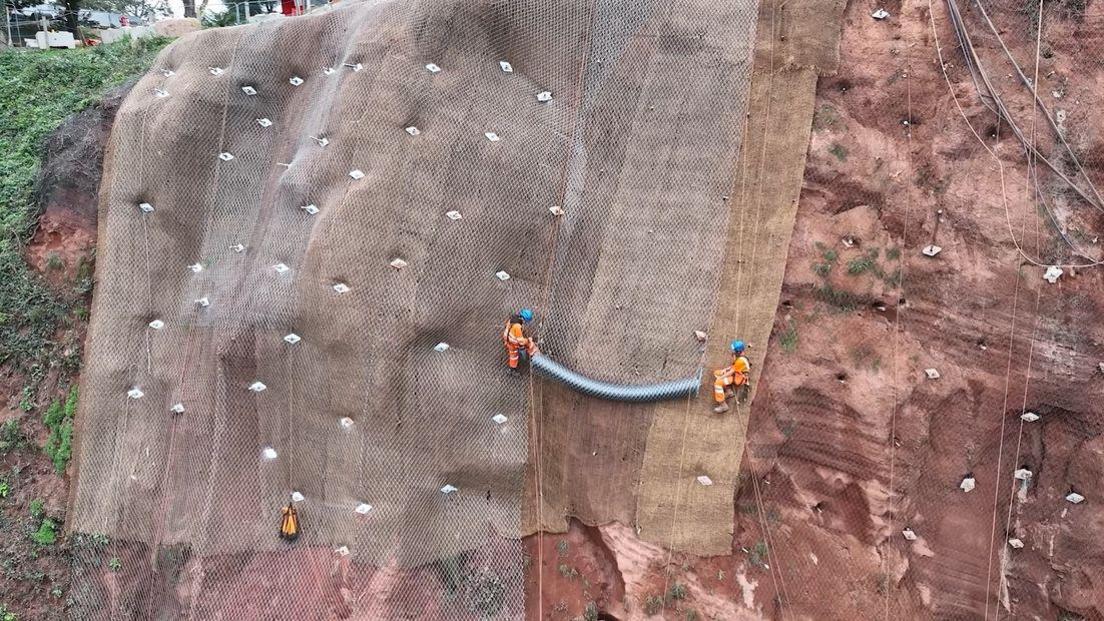 Stainless steel netting being secured to the cliffs above the railway line. Two works in orange uniforms are seen unravelling the netting.