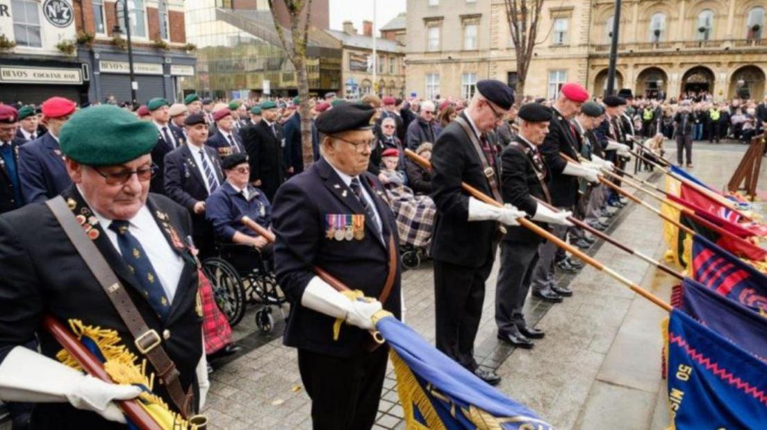 A crowd of men holding flags that are laid down in front of them and army uniforms. Members of the public can be seen in the distance with two police officers dotted in front of them. 