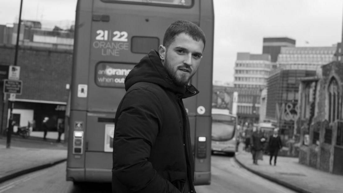 A black and white photo of a young man which short hair and a beard. He is wearing a thick black coat and is stood in the middle of a road with a double decker bus and a busy street in the background. 