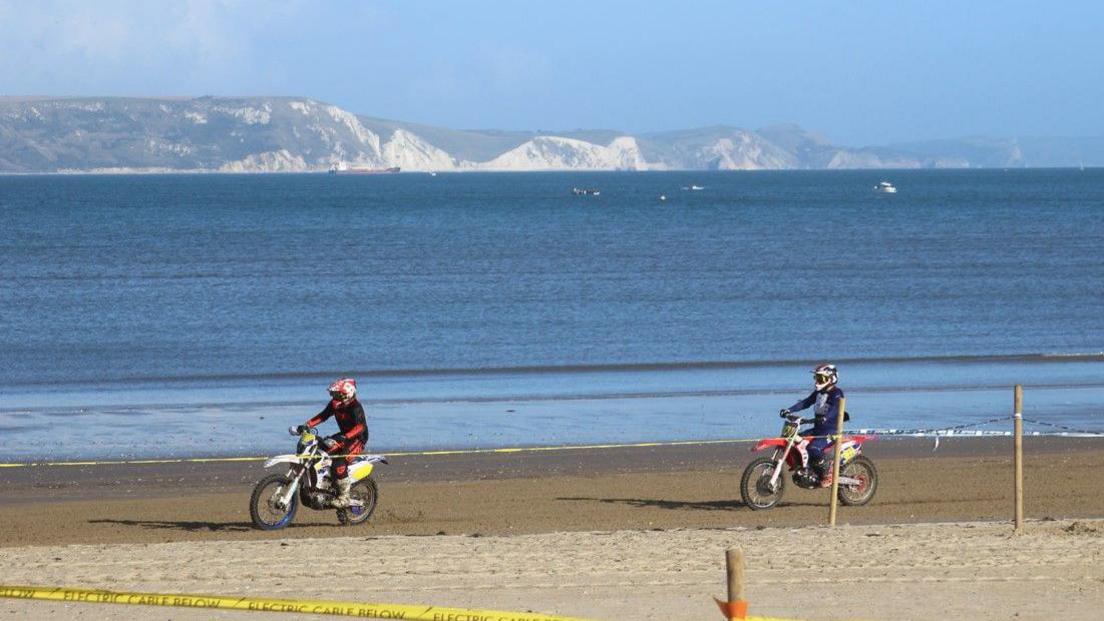 Two motocross riders on their bikes on a firm part of the beach track nearest the water. The white cliffs on the other side of the bay are in the background