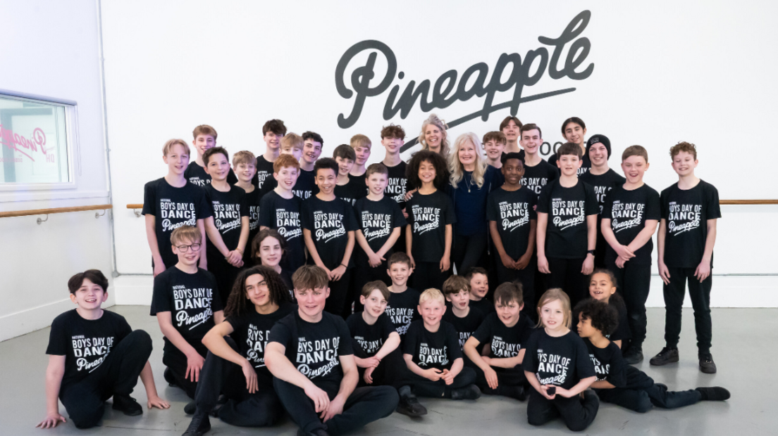 40 male dancers all looking at the camera and wearing black dance trousers and t-shirts with white writing on. Some of the team are standing, whilst others are sitting on the floor all in front of a white wall with a black Pineapple logo.