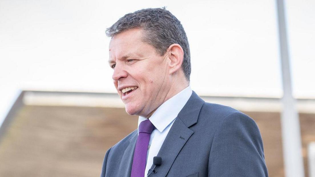 Rhun ap Iorwerth speaking in front of the Senedd building, wearing a light blue suit and purple tie, with a microphone on his lapel.