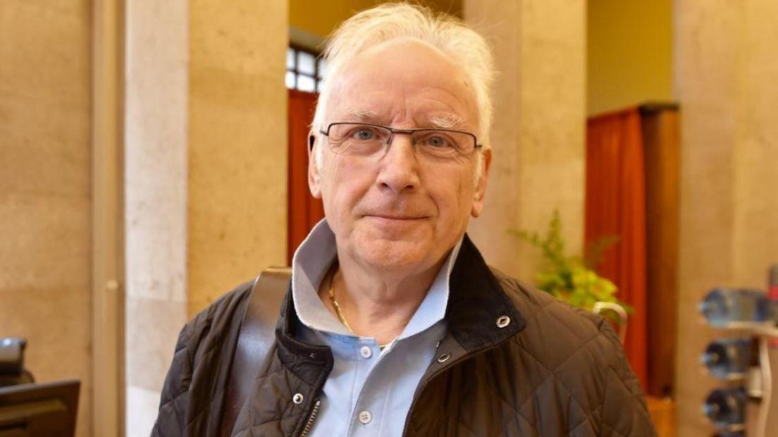 Pete Waterman standing in the lobby of a building. He has an open neck shirt and is wearing a black jacket. He is wearing glasses, a gold chain and has a slight smile. 