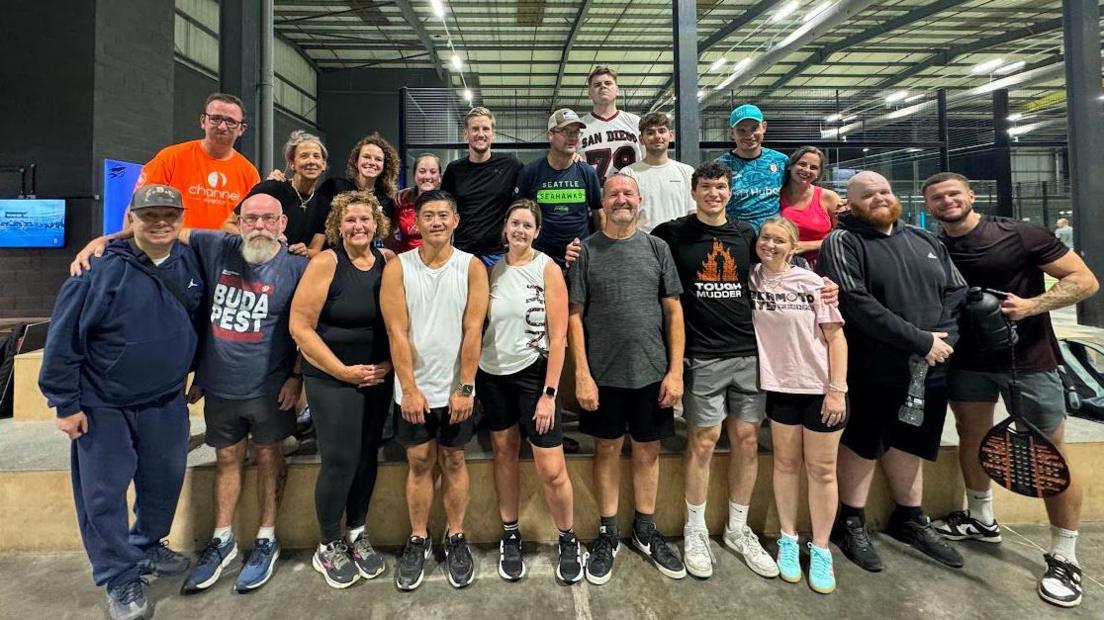 A large diverse group of people smiling at the camera at the Rocket Padel indoor centre. They are all dressed in sports gear and some are posing with rackets.