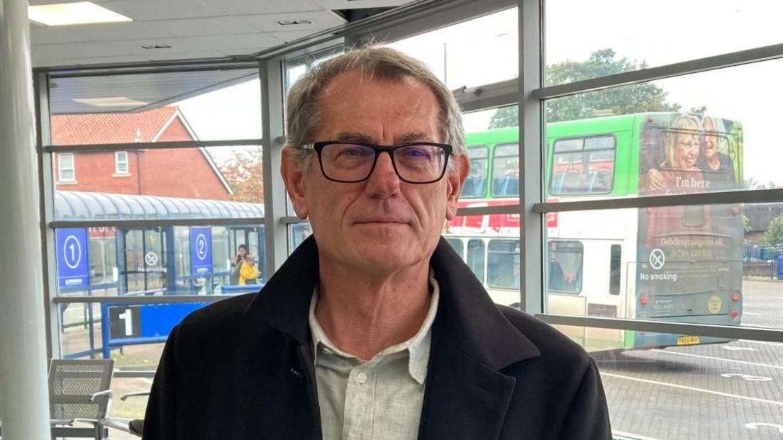 A man wearing a dark coat and a light shirt standing in a bus shelter. Behind him is a bus.