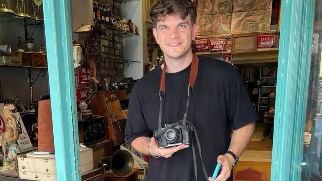 Conor Buckett is standing in the doorway to an antiques shop with an old camera strung around his neck. He is wearing a black t-shirt and staring at the camera.