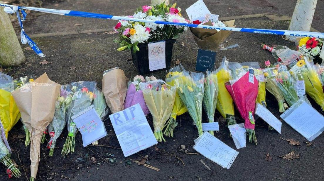 At least 16 bunches of flowers on the ground. Police tape is higher up on the photo. The tape is stretched between concrete bollards.