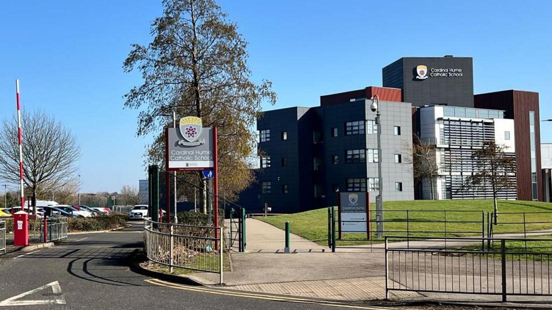 View of the front entrance to Cardinal Hume Catholic School. On the left is the road into the car park and the school building on the right. It is a modern grey of three storeys with signage and a grassed area in front.