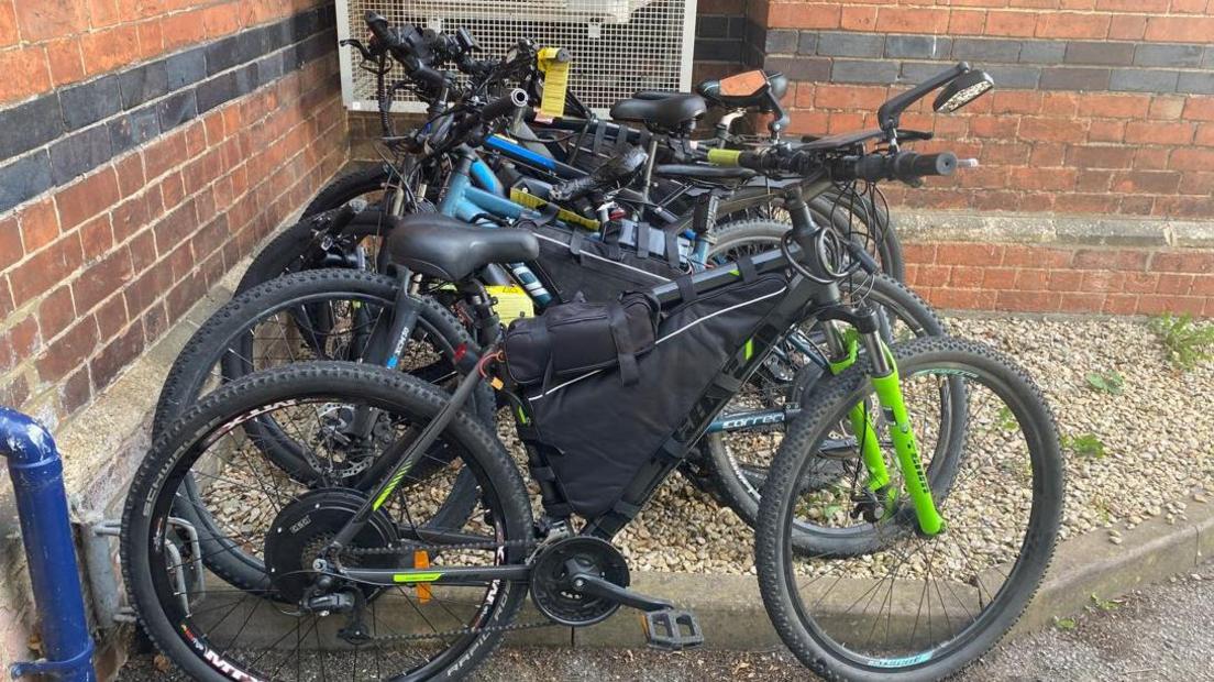 A row of at least five bicycles parked against a brick wall. They have different accessories including bags, locks and large batteries.