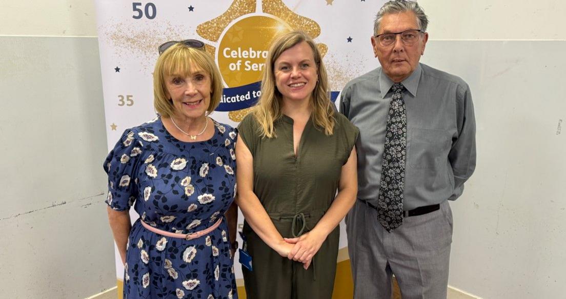Maureen Lavin (left) has shoulder-length fair hair and is wearing a blue dress with white flowers on it. Laura Churchward is standing in the centre and has long fair hair and is wearing a dark green dress. David Knight (right), has short grey hair and is wearing spectacles. He has a grey shirt on and grey trousers and a pattered dark tie