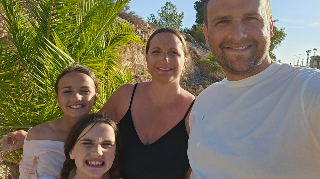 Alexander David wearing a white t-shirt smiling at the camera. He is stood with a woman, and his two young daughters.