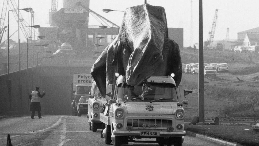 A model dinosaur on top of a car being taken across the Dartford Crossing