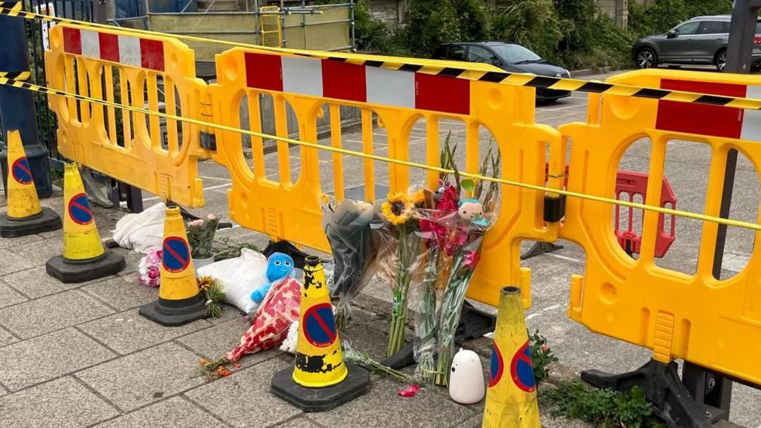 Flowers and a blue cuddly toy attached to a yellow temporary road barrier