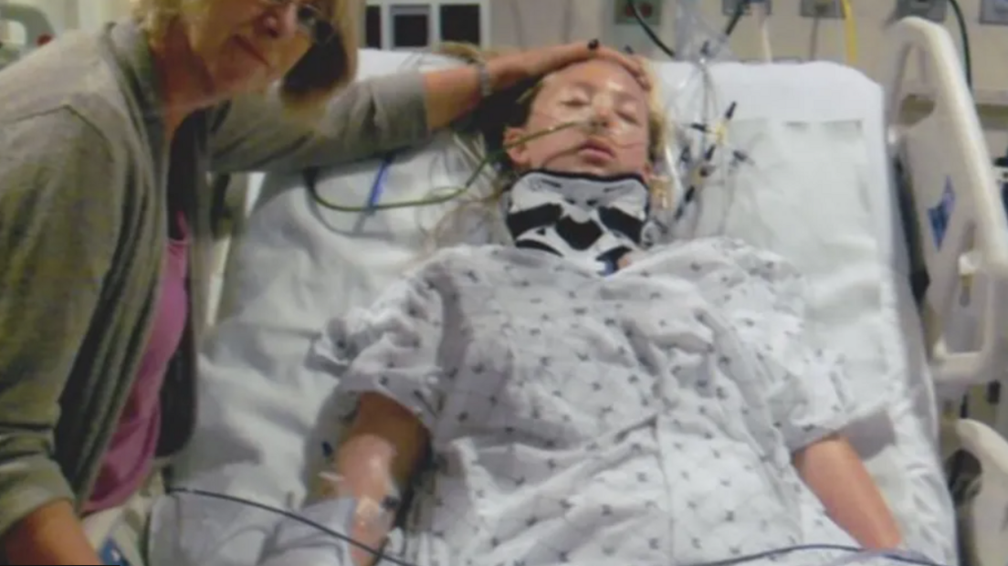 A teenage Sophie Carrigill lies in a hospital bed with a neck brace on and various tubes attached to her, next to her is a woman in a grey cardigan who is touching her head