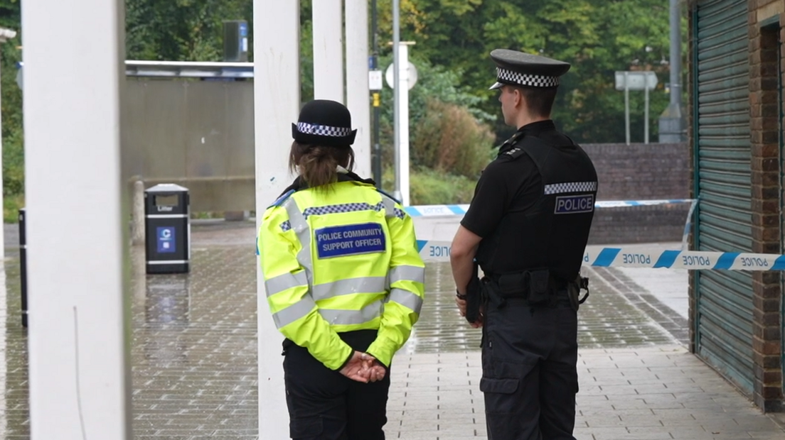 One female and one male police office standing by a police cordon. The female officer had a hat and yellow high vis jacket on with her arms  behind her back, facing away from the camera. The male officer is wearing all black, a hat, and is also facing away from the camera.