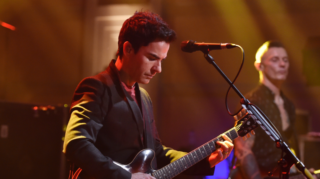 Lead singer Kelly Jones is playing guitar in front of a microphone. He has short black hair and is wearing a suit jacket.