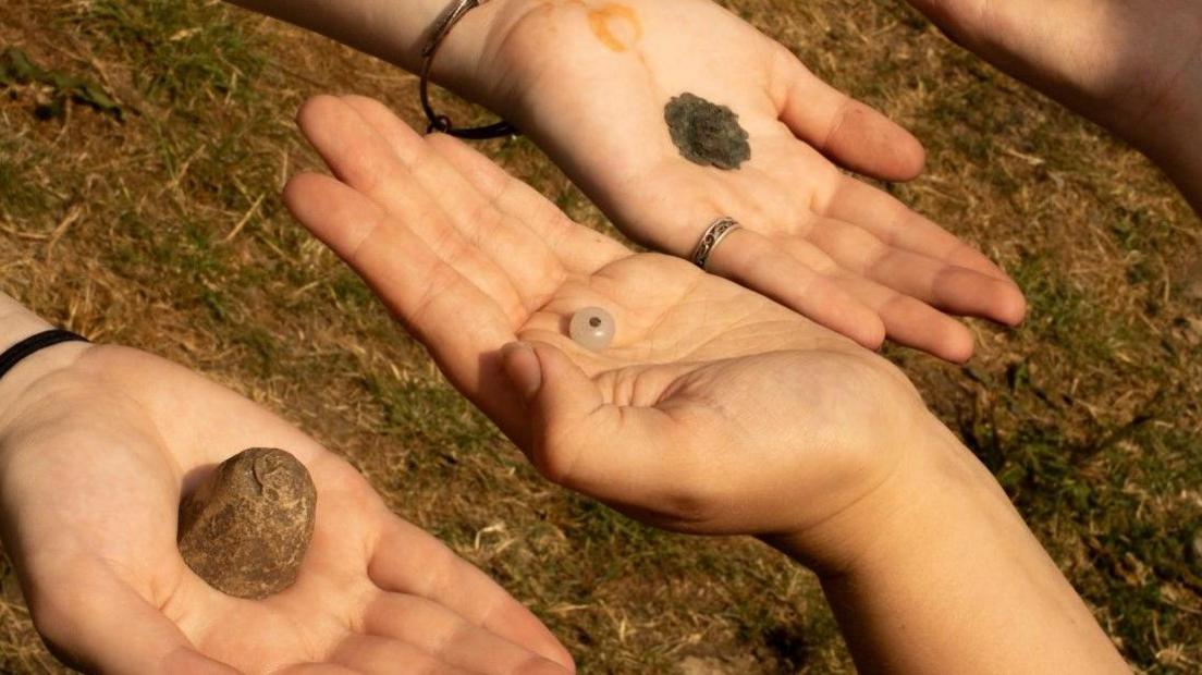 Three palms stretching out with objects found during the excavation placed on top of them, including two metal pieces and a bead.