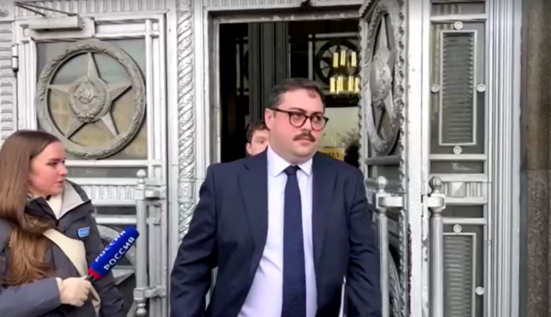 A man, dressed in a suit with a blue tie and wearing dark-rimed glasses, leaves building with ornately decorated doors