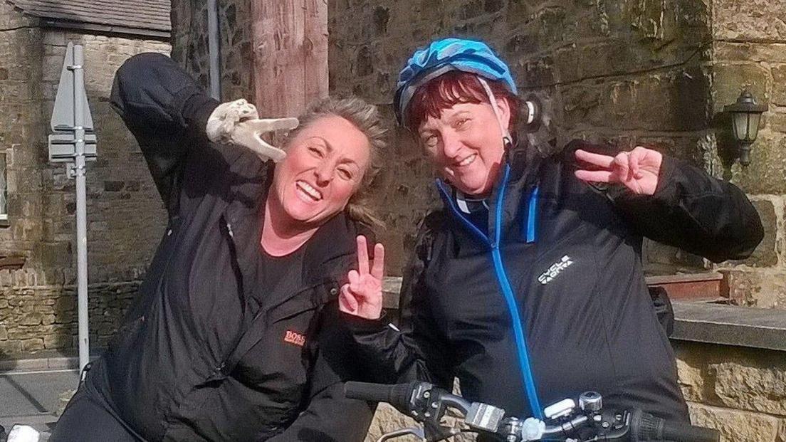 Debbie Bailey and her sister Tracey Bailey on bikes in the Peak District. They are both smiling and holding up peace signs. 