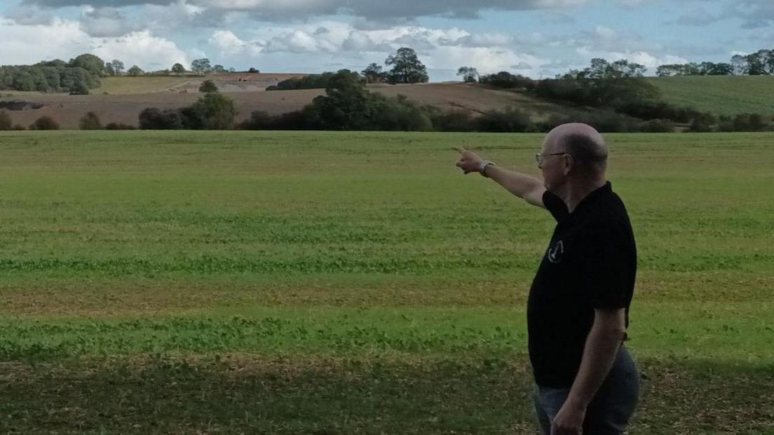 Graham Evans in a black shirt standing in a field pointing out where a soil storage site is located on part of a historic battlefield. The battlefield is large rolling fields. 
