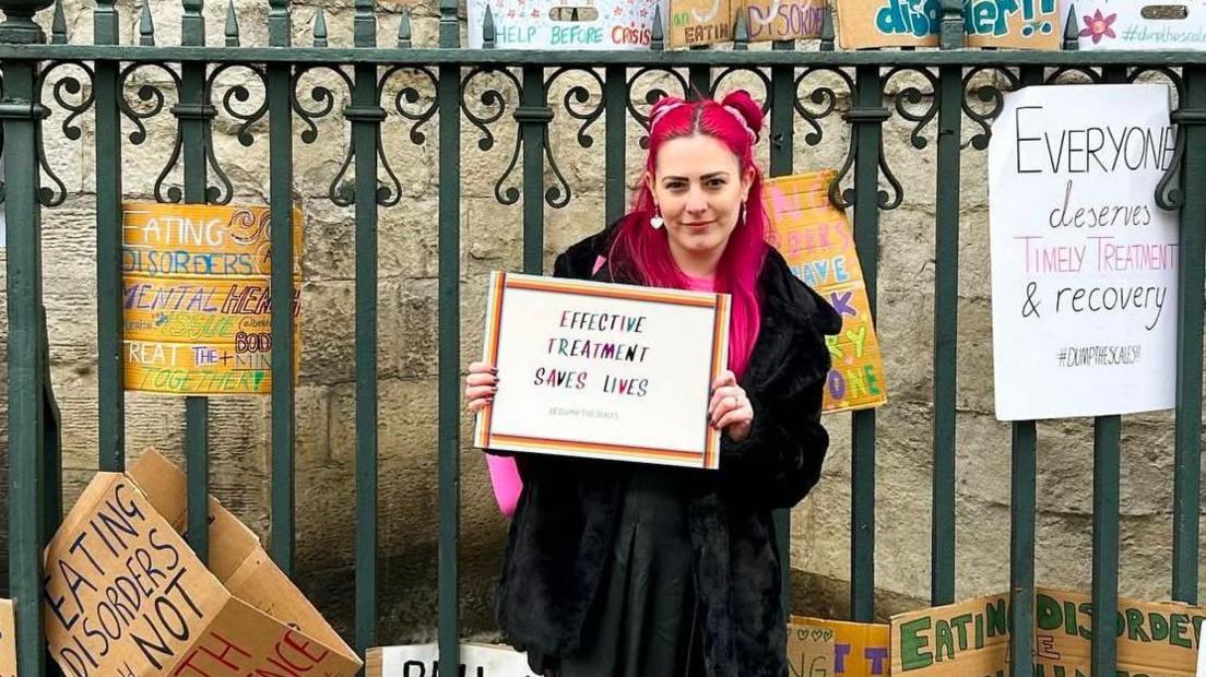 A woman with pink hair in front of a green metal fence holding a sign that says EFFECTIVE TREATMENT SAVES LIVES