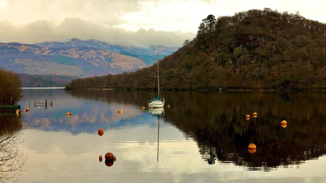 A boat moored in the middle of Loch Lomond. The boat is white. It has a mast reaching half way up the picture. It is sitting on still water surrounded by orange buoys. There are mountains in the background. 