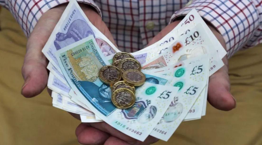 A man in a white shirt with blue and red checks holds out his hands, on which is a pile of cash. Twenty, ten and five pounds notes are fanned in his hands and seven pound coins placed a top the notes. 