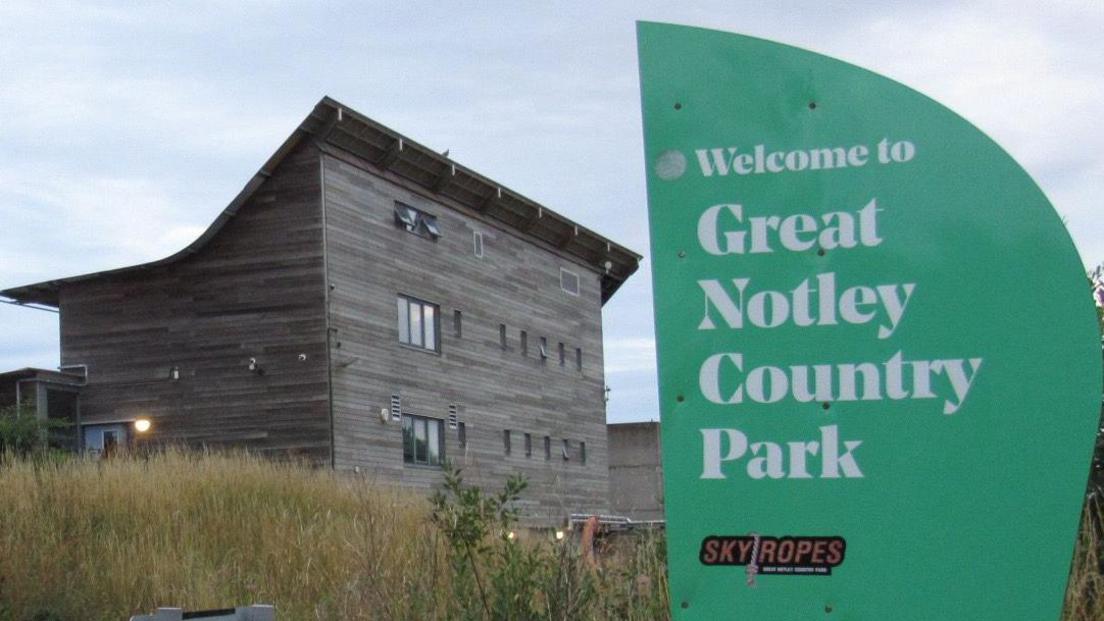 A large building made of long wooden panels. It is three storeys high at the front, with a sloping, curved roof to the back, which has two storeys. In the foreground is a green sign that says "Welcome to Great Notley Country Park" in white font.
