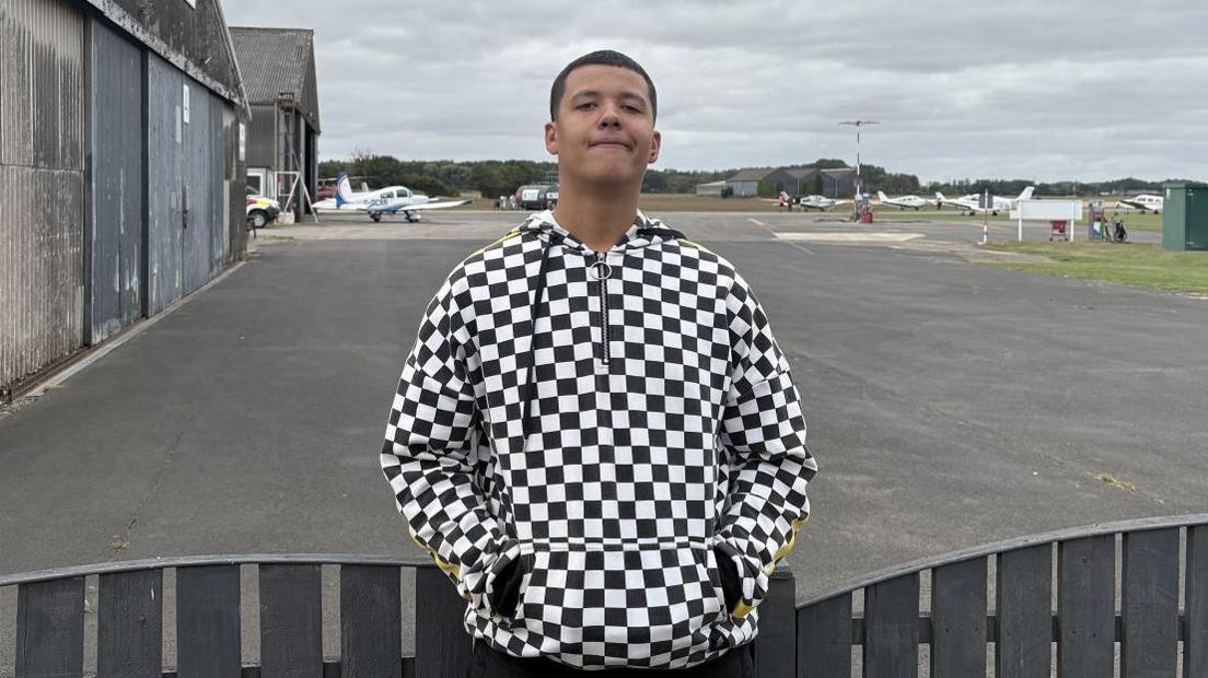 A teenage boy stands by a wooden fence on an airfield with prop planes in the background. He wears a black and white chequered coat.