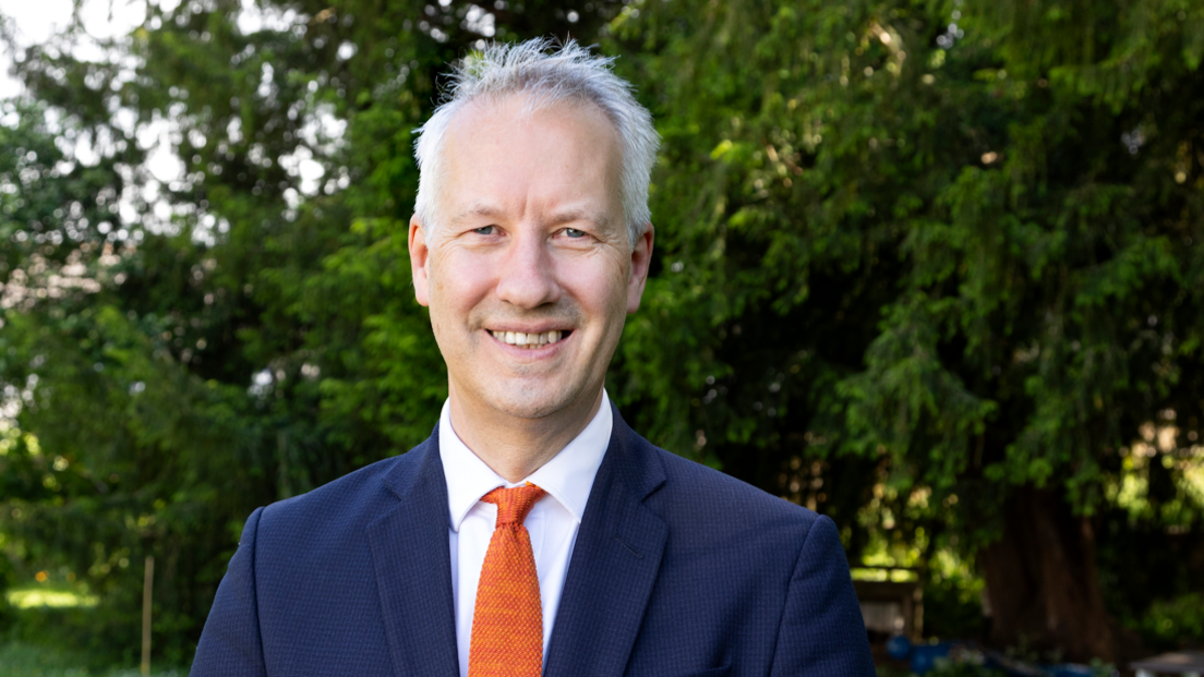 Gideon Amos looking at a camera with green trees behind him