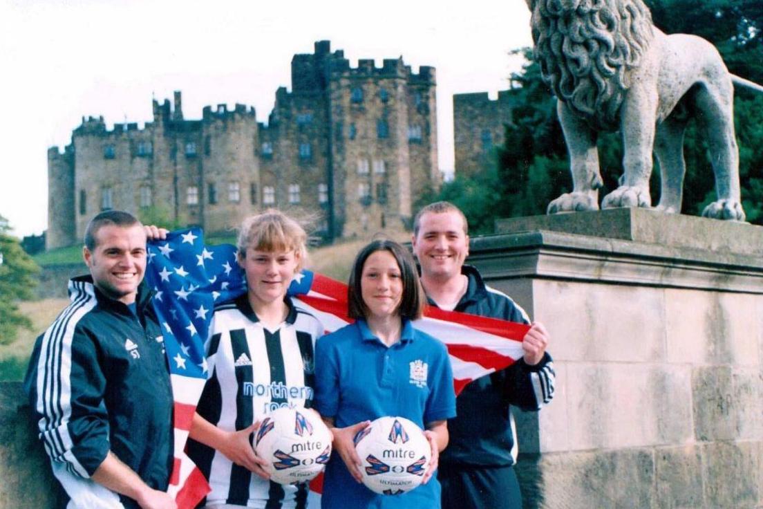 A young Lucy Bronze holding a football as she stands next to fellow player Danni Pope and coaches Shaun and Ryan Jobson. Alnwick Castle can be seen in the background while a stone lion is to their right.