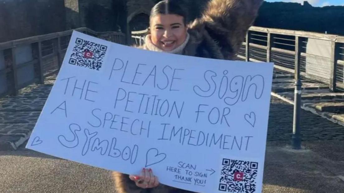 Jessie Yendle holding white placard with QR codes and message saying please sign the petition for a speech impediment symbol