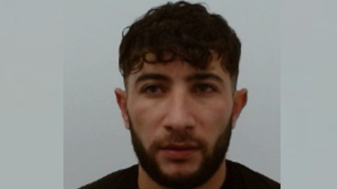 a police mugshot of Muhammad Sheikhi who has short, black hair and a black beard. He is wearing a black T-shirt.