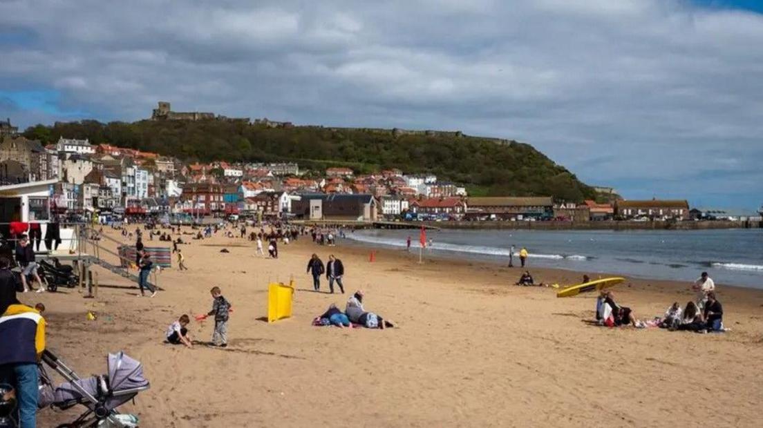 It is a busy, sunny day on Scarborough's South Bay beach but there a dark clouds above. People are lying and sitting on the sand in the foreground, the donkeys are giving people rides in the distance.