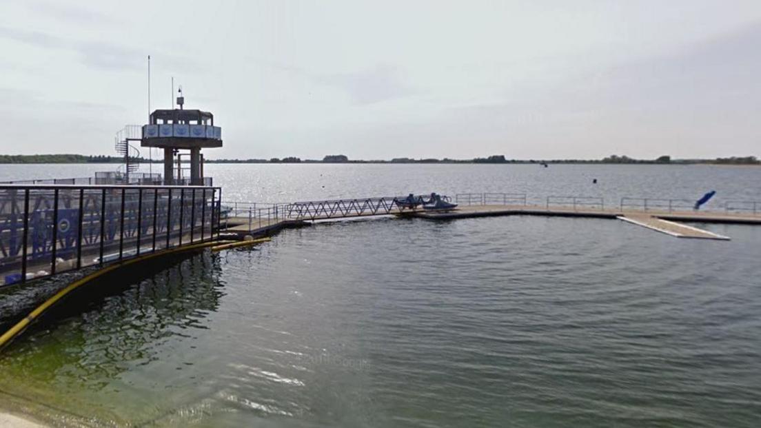 Farmoor Reservoir with a large jetty and lookout point pictured in the foreground. It is constructed of a metal framework and floating pontoons. The water is greenish blue. Trees on the other side of the reservoir are visible in the distance.