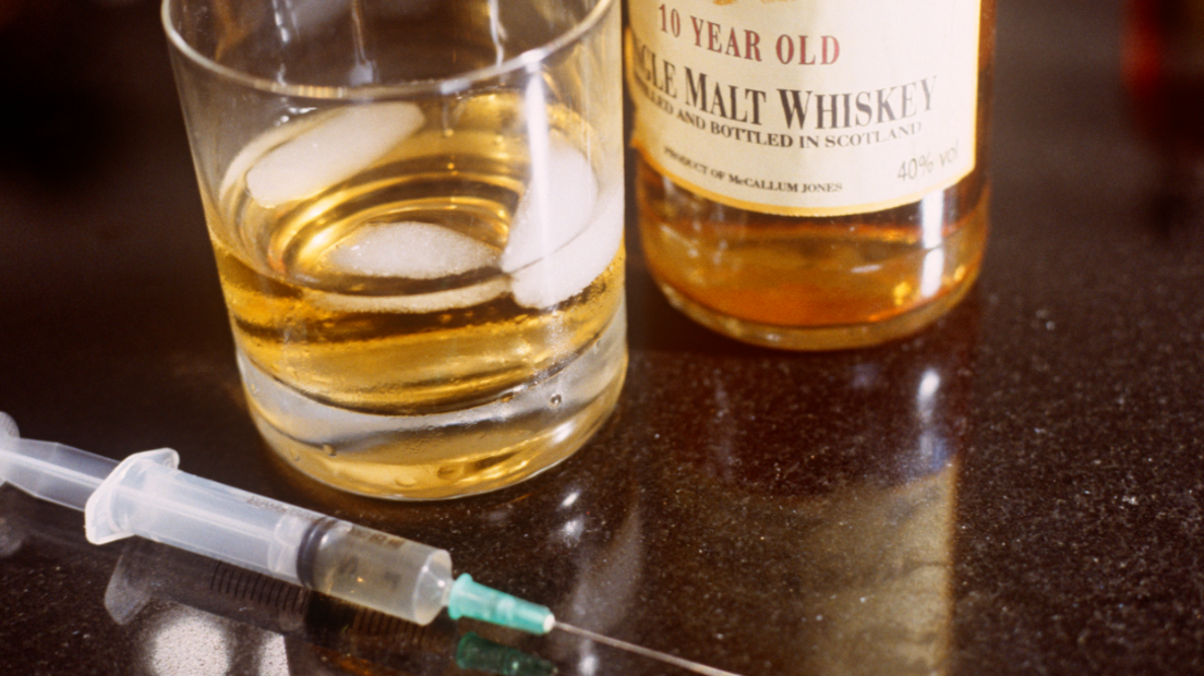 A bottle of whiskey, a glass with whiskey and ice, and a syringe sitting on a table 