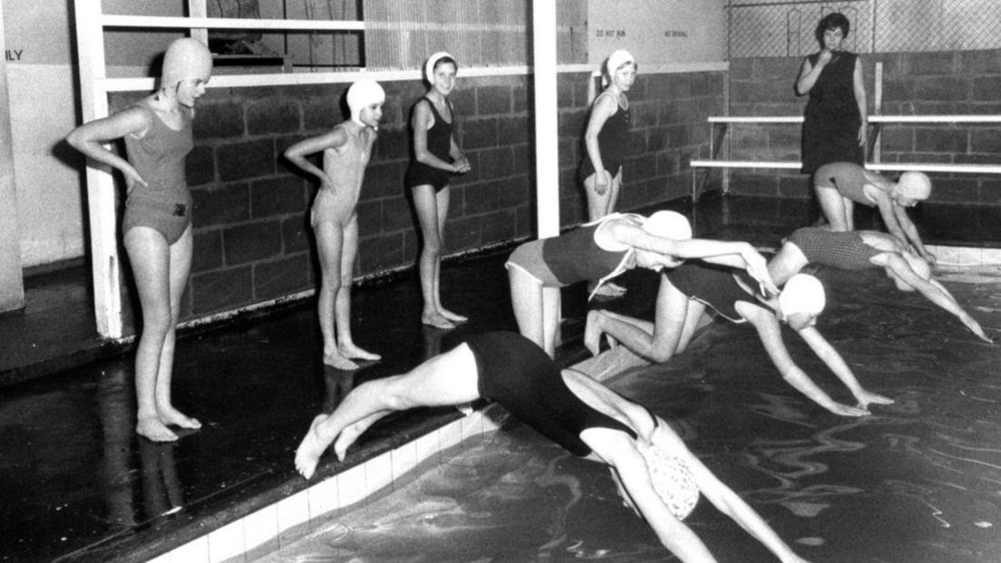 A black and white photo of a group of nine young girls diving into a pool or wait to dive in. A female teacher can be seen in the background blowing a whistle. The walls of the building seem to be breeze block.