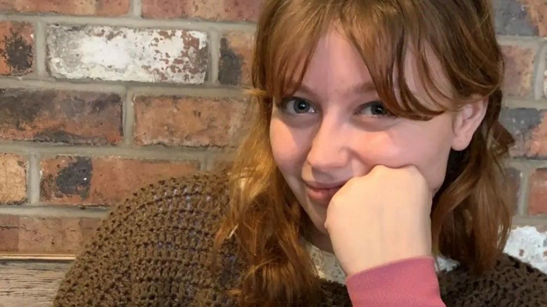 Kat Nellist is looking at the camera while resting her face against her closed fist while her elbow rests on the table. She has shoulder-length wavy auburn hair and curtain bangs. She is wearing a brown knitted jumper and is sitting in front of a brick wall