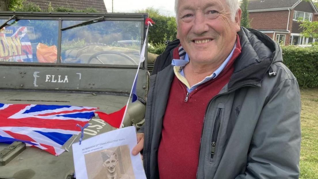 A man wearing a grey coat and red fleece is smiling at the camera and holding a picture of his father. An old green vehicle is behind him.