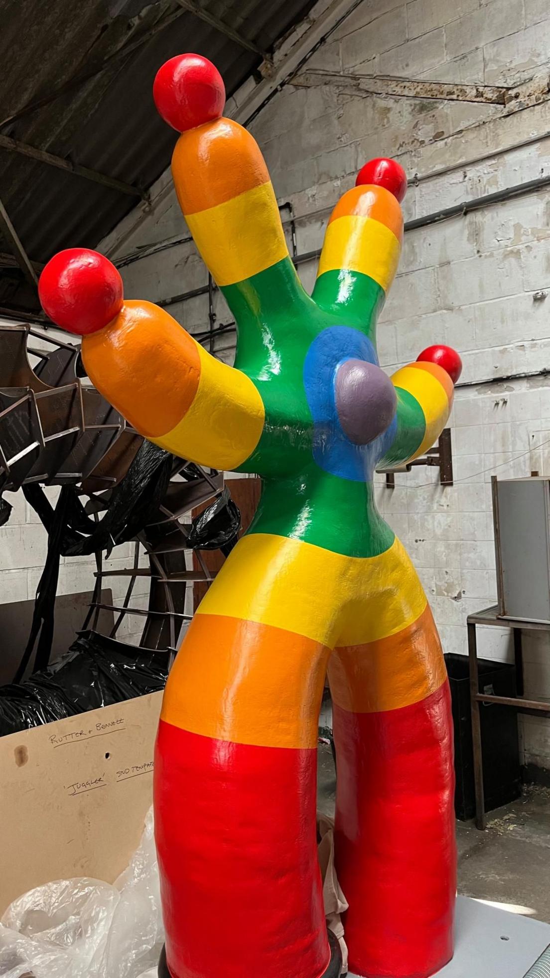 A four-armed, two legged sculpture painted in concentric bands (from outer to inner) red, orange, yellow, green, blue and purple. The design has a ball in the middle and on the tips of all four arms. The sculpture stands in a warehouse.