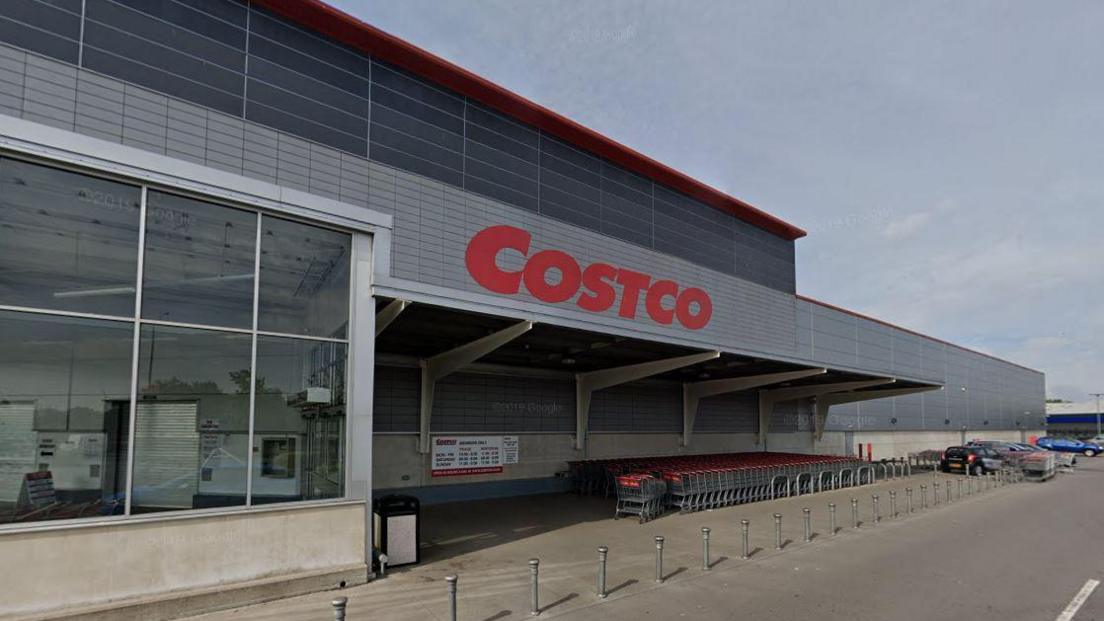 A screenshot from Google street view showing the exterior entrance to Costco. It is a large grey warehouse building with the store's name in red across the front and a large window on the left. Under a sheltered area there are lots of trolleys lines up. To the right there is a vast car park. 