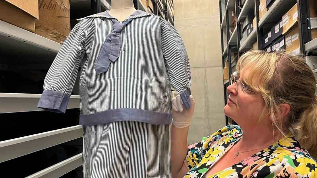 A woman wearing glasses and a floral dress examines a children's sailor suit within a museum storage area. The suit has blue and white pin-stripes, medium-blue trimmings and a blue tie.