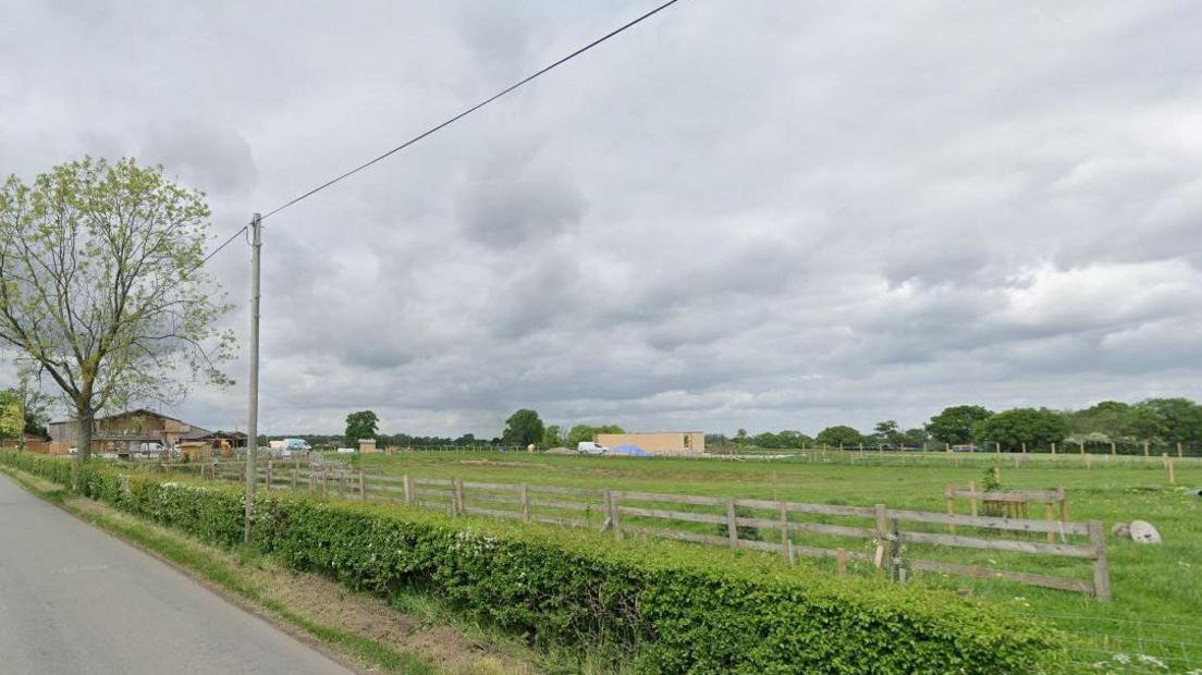 A view of a field from a country lane. There is a line of trees in the distance and, to the left of the shot in the background, the dairy farm near to which the zoo would be built. 