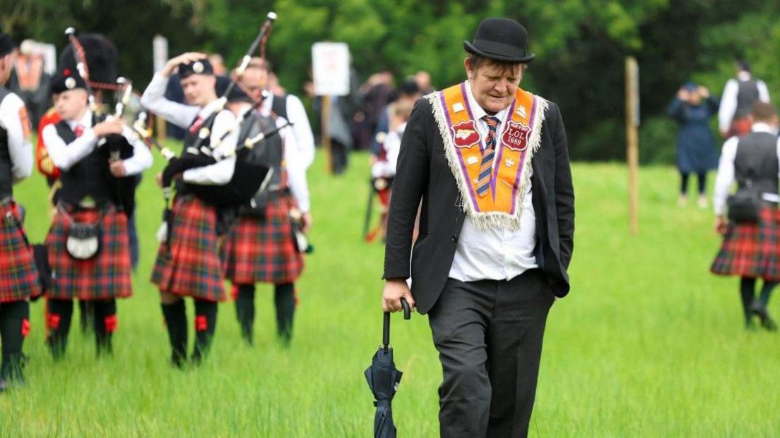 A man is walking in a field with an umbrella in his right hand. He is looking down at the ground, wearing a dark suit, black bowler hat and an orange sash around his shoulders.