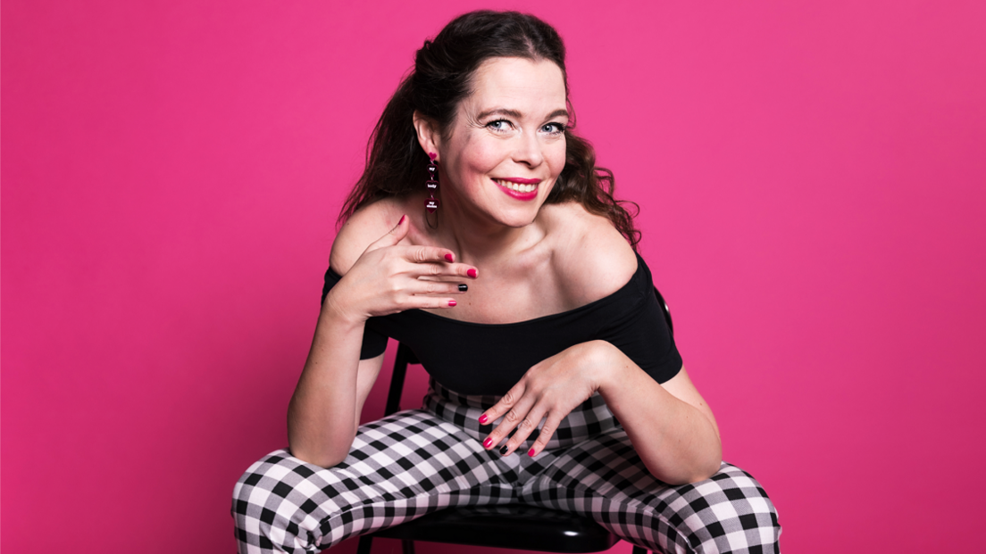 Juliette Burton sitting on a black chair wearing black gingham trousers and a black off-the-shoulder top, posing in front of a shocking pink background