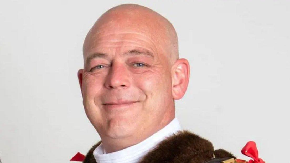 A headshot photo of a man wearing mayoral clothes. He has a bald head and is smiling at the camera.